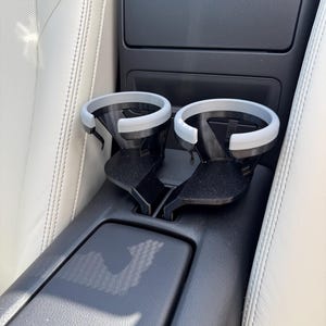 Puede incluir: Un portavasos negro para coche con dos aberturas circulares, cada una con un borde gris claro. El portavasos está diseñado para encajar en la consola central de un coche, como se ve en la imagen.