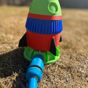 May include: A colorful rocket-shaped lawn sprinkler. The sprinkler features a red body, green top, blue accents, and black fins. A blue hose is connected to the base. The sprinkler is designed for outdoor use.