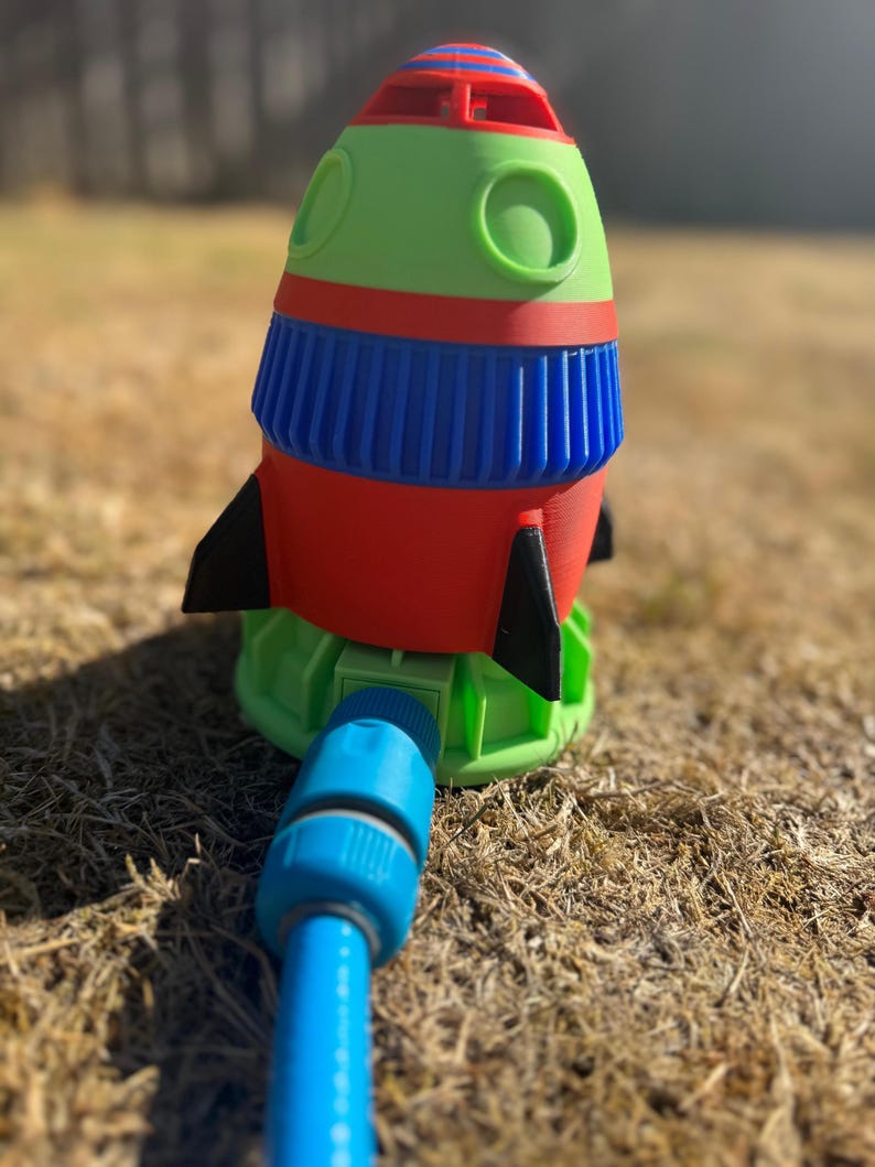 May include: A colorful rocket-shaped lawn sprinkler. The sprinkler features a red body, green top, blue accents, and black fins. A blue hose is connected to the base. The sprinkler is designed for outdoor use.