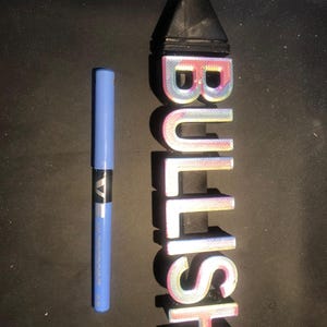 May include: A 3D printed sign that says "BULLISH" in iridescent pink, blue, and white letters. The sign is about 25 cm tall and is sitting on a black surface.