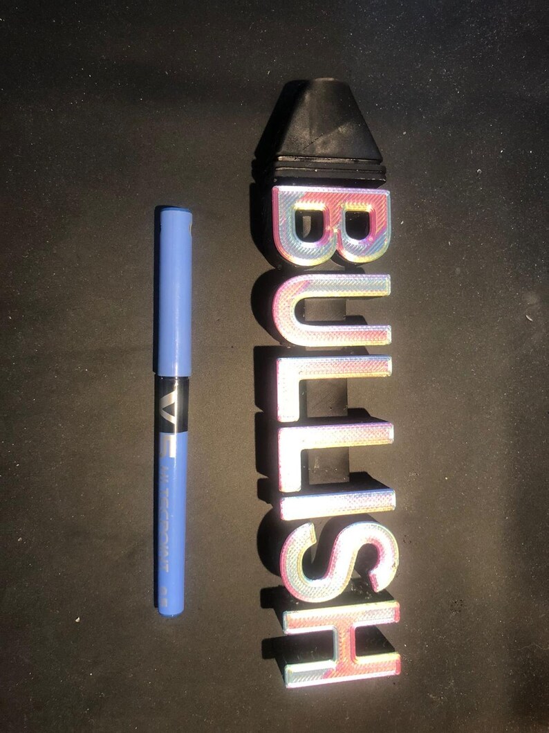 May include: A 3D printed sign that says "BULLISH" in iridescent pink, blue, and white letters. The sign is about 25 cm tall and is sitting on a black surface.