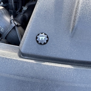 May include: Close-up of a black car engine component. A small, circular cap with the letters "TT" in white is centered on a textured, dark gray surface. The cap has a gear-like edge.