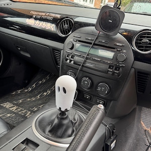 Op de afbeelding: Auto-interieur met een witte spookvormige versnellingspook. Het zwarte dashboard heeft een draadloze oplaadpad en de tekst "EVERYTHING IS CHROME IN THE FUTURE!". Het interieur van de auto is zwart.