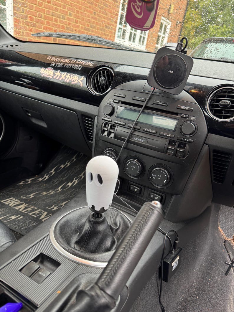 Op de afbeelding: Auto-interieur met een witte spookvormige versnellingspook. Het zwarte dashboard heeft een draadloze oplaadpad en de tekst "EVERYTHING IS CHROME IN THE FUTURE!". Het interieur van de auto is zwart.