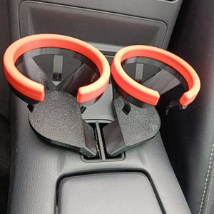 May include: A pair of black car cup holders with bright orange rims. The cup holders are designed to fit into a car's center console. The image shows the cup holders installed in a car, with a focus on their design and color.