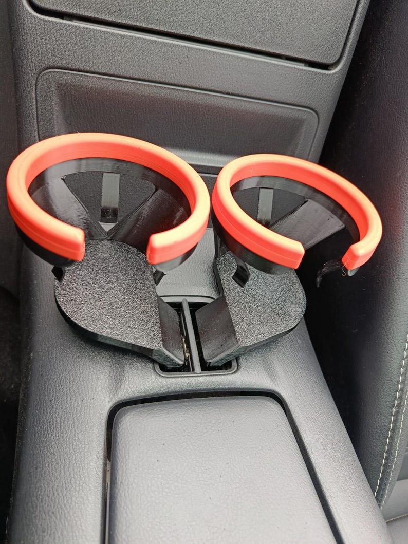 May include: A pair of black car cup holders with bright orange rims. The cup holders are designed to fit into a car's center console. The image shows the cup holders installed in a car, with a focus on their design and color.
