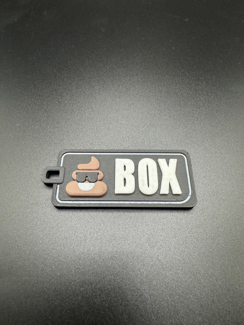 May include: A black and white patch with a brown poop emoji wearing sunglasses and the word "BOX" in white letters.