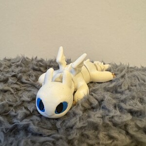May include: White 3D printed dragon figurine with blue eyes, lying on a gray fluffy surface.