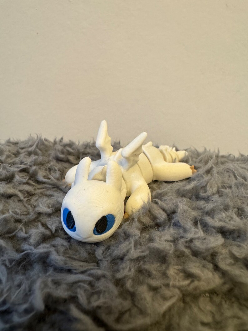 May include: White 3D printed dragon figurine with blue eyes, lying on a gray fluffy surface.