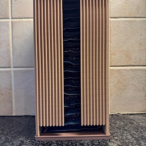May include: A vertical, rectangular coffee pod holder made of light brown wood. The front features vertical slats that slide open to reveal the coffee pods inside. The holder is designed to sit on a countertop and organize coffee pods.