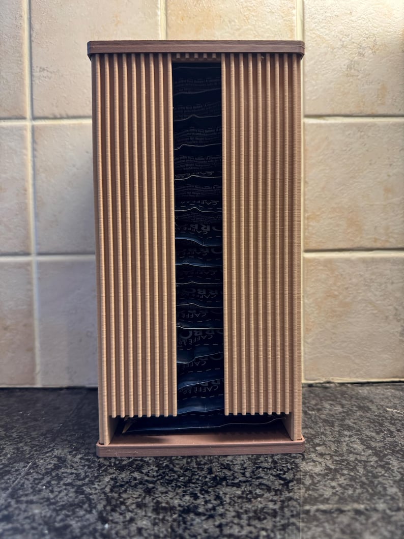 May include: A vertical, rectangular coffee pod holder made of light brown wood. The front features vertical slats that slide open to reveal the coffee pods inside. The holder is designed to sit on a countertop and organize coffee pods.