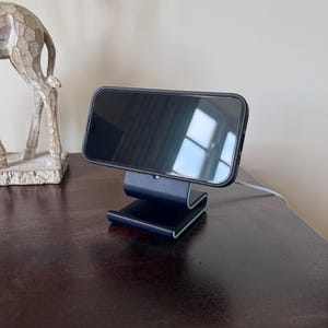 May include: A black smartphone rests on a dark blue phone stand with a curved design. The stand has a light blue accent on the edges. A decorative silver giraffe statue is visible in the background.