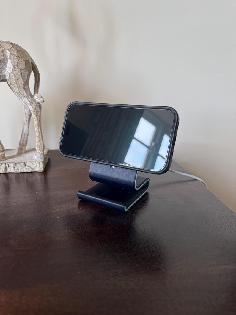 May include: A black smartphone rests on a dark blue phone stand with a curved design. The stand has a light blue accent on the edges. A decorative silver giraffe statue is visible in the background.