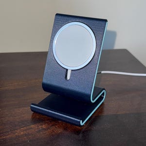 May include: A dark blue phone charging stand with a white circular charging pad. The stand has a curved design with light blue accents. A white charging cable is connected to the stand.