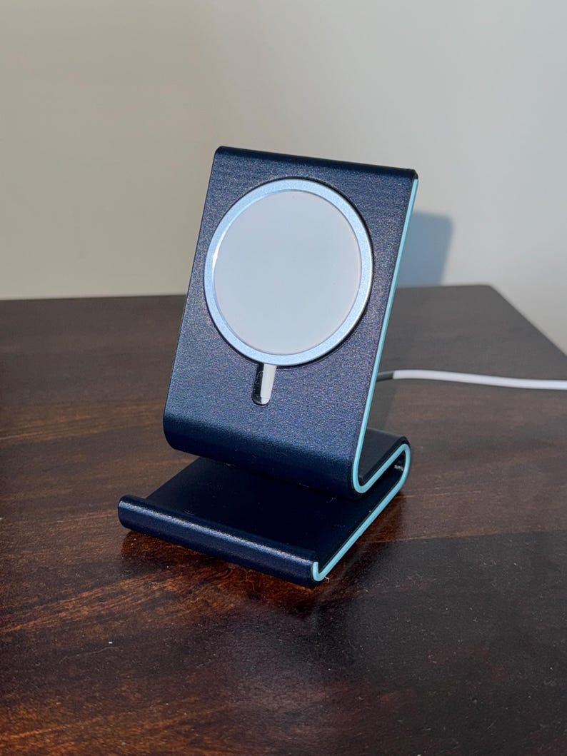 May include: A dark blue phone charging stand with a white circular charging pad. The stand has a curved design with light blue accents. A white charging cable is connected to the stand.