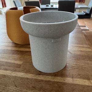 May include: A light gray speckled planter with a wide, shallow top and a cylindrical base. An orange textured vase is to the left. The objects are on a wooden table. The background includes a dining room with chairs and a window.