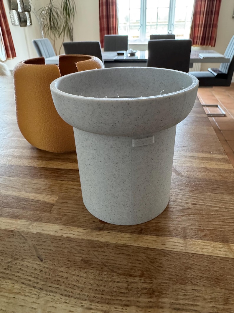 May include: A light gray speckled planter with a wide, shallow top and a cylindrical base. An orange textured vase is to the left. The objects are on a wooden table. The background includes a dining room with chairs and a window.