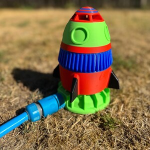 May include: A colorful rocket-shaped lawn sprinkler. The sprinkler features a red, green, and blue design with black fins. A blue hose is attached to the base, ready for watering the lawn. The sprinkler is sitting on dry grass.