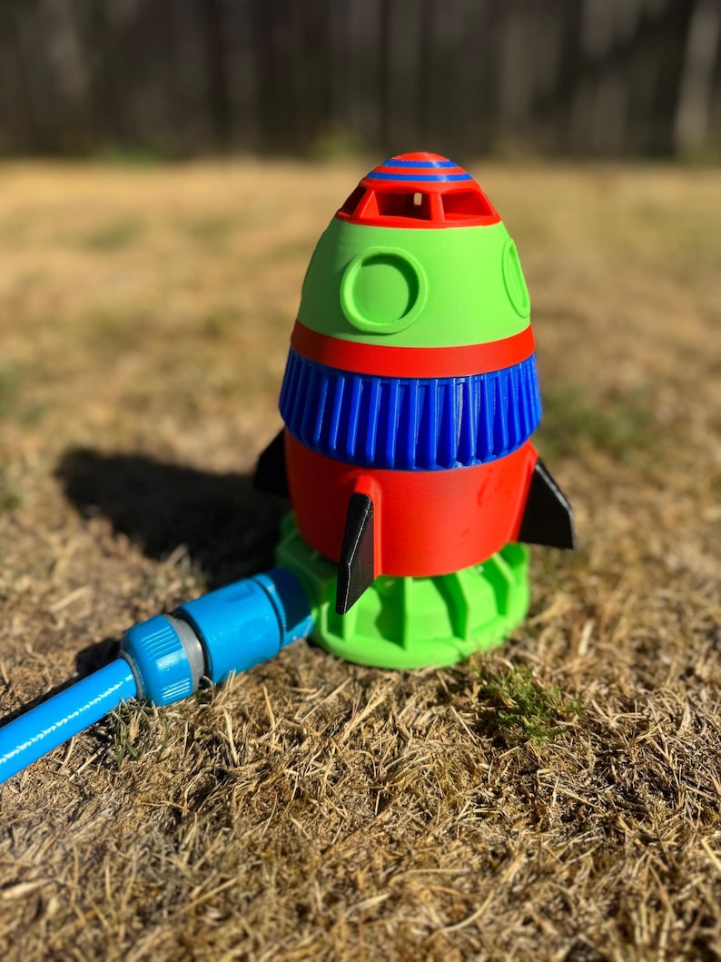 May include: A colorful rocket-shaped lawn sprinkler. The sprinkler features a red, green, and blue design with black fins. A blue hose is attached to the base, ready for watering the lawn. The sprinkler is sitting on dry grass.