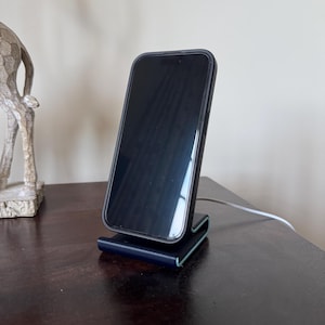May include: A black smartphone in a black case is propped up on a dark blue charging stand. The stand has a curved design with a light green edge. A white charging cable is connected to the stand.