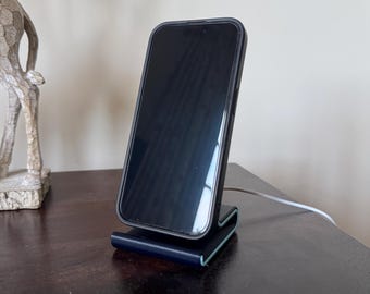 Op de afbeelding: Een zwarte smartphone in een zwarte hoes staat op een donkerblauwe oplaadstandaard. De standaard heeft een gebogen ontwerp met een lichtgroene rand. Een witte oplaadkabel is aangesloten.