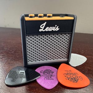 May include: A black and orange miniature guitar amplifier with the word "Lewis" printed on the front. The amplifier is sitting on a wooden surface with four guitar picks in front of it.
