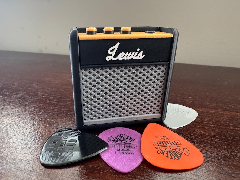 May include: A black and orange miniature guitar amplifier with the word "Lewis" printed on the front. The amplifier is sitting on a wooden surface with four guitar picks in front of it.