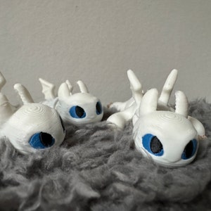 May include: Three white plush toys shaped like dragons with blue eyes. The toys are lying on a gray fluffy surface.
