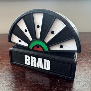 May include: A black and white dartboard with a green and red bullseye. The dartboard is mounted on a black base with the name "BRAD" printed in white letters.