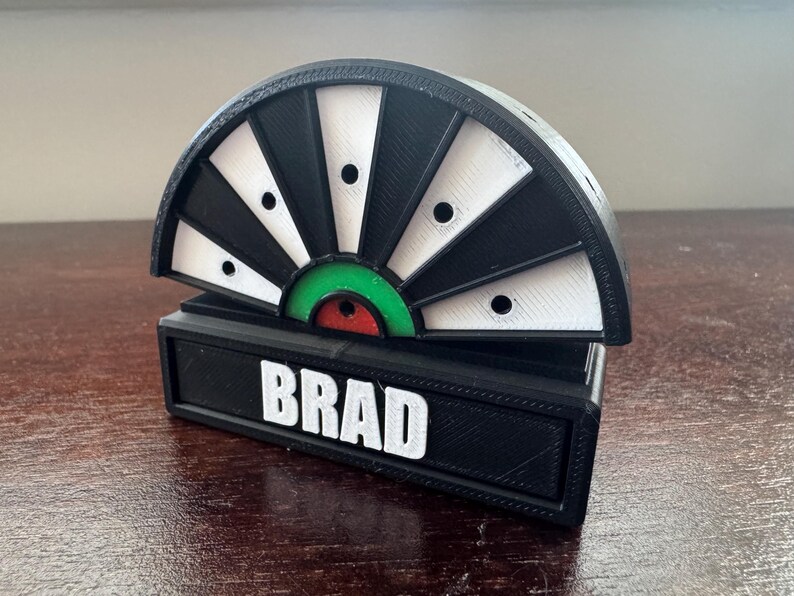 May include: A black and white dartboard with a green and red bullseye. The dartboard is mounted on a black base with the name "BRAD" printed in white letters.