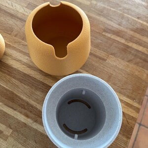May include: Two-piece, round, textured containers. The top container is a light orange with a curved opening. The bottom container is light gray with two curved cutouts inside. The containers are on a wooden surface.