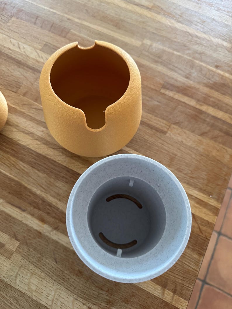 May include: Two-piece, round, textured containers. The top container is a light orange with a curved opening. The bottom container is light gray with two curved cutouts inside. The containers are on a wooden surface.