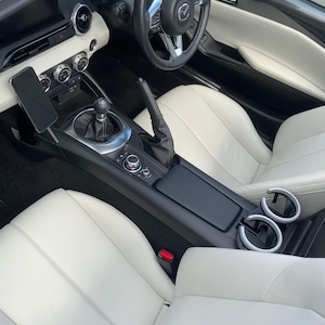 May include: Interior view of a black convertible car with white leather seats. The steering wheel is black with silver accents. The center console features a gear shift, cup holders, and a phone mount. The seats have a stitched design.