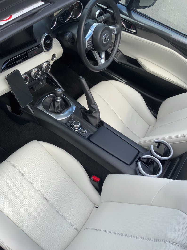 May include: Interior view of a black convertible car with white leather seats. The steering wheel is black with silver accents. The center console features a gear shift, cup holders, and a phone mount. The seats have a stitched design.