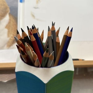 May include: A blue and green pencil holder shaped like a pencil, filled with a variety of coloured pencils.