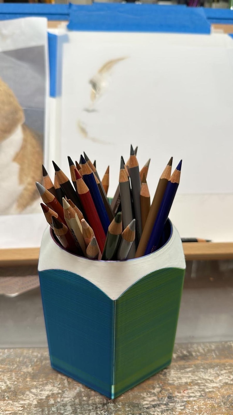 May include: A blue and green pencil holder shaped like a pencil, filled with a variety of coloured pencils.