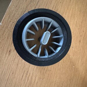 May include: A gray and black 3D-printed wheel-shaped object. The wheel has a central hub with spokes radiating outwards. The outer rim is black, and the spokes and hub are gray. The object is set into a wooden surface.