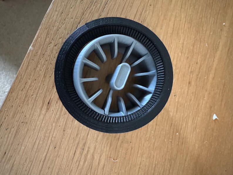 May include: A gray and black 3D-printed wheel-shaped object. The wheel has a central hub with spokes radiating outwards. The outer rim is black, and the spokes and hub are gray. The object is set into a wooden surface.