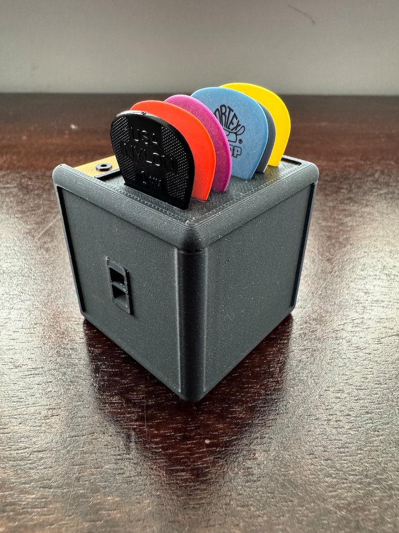 May include: A black plastic guitar pick holder shaped like a small amplifier with a variety of colored guitar picks inside.