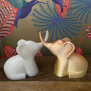 Op de afbeelding: Twee kleine olifantenfiguurtjes, een witte en een gouden, staan ​​tegenover elkaar met hun slurfjes die elkaar raken. De figuurtjes staan ​​op een houten oppervlak met een achtergrond van een kleurrijk bloemenpatroon.