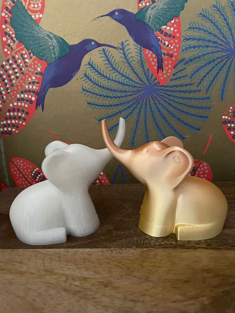 Op de afbeelding: Twee kleine olifantenfiguurtjes, een witte en een gouden, staan ​​tegenover elkaar met hun slurfjes die elkaar raken. De figuurtjes staan ​​op een houten oppervlak met een achtergrond van een kleurrijk bloemenpatroon.