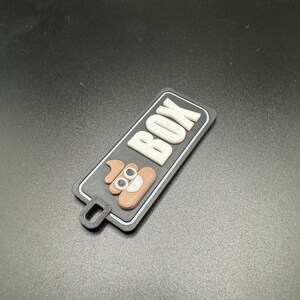 May include: A black and white rectangular tag with the word "POOP BOX" in white letters. A brown poop emoji with white eyes is in the centre of the tag.