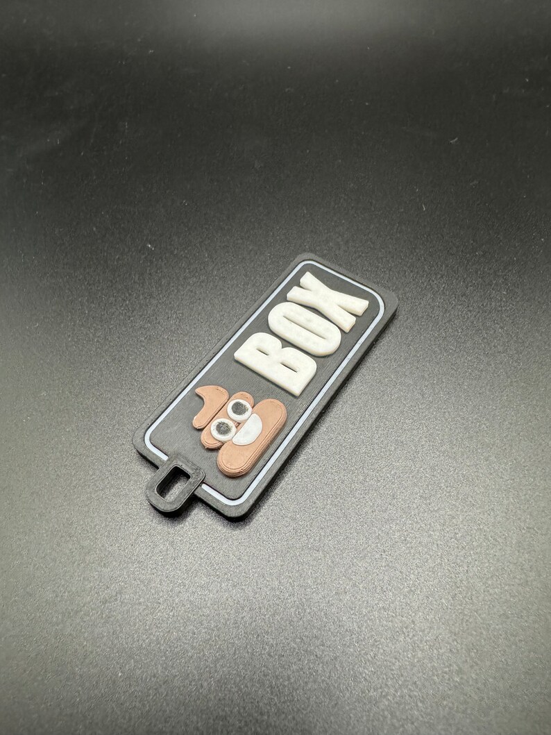 May include: A black and white rectangular tag with the word "POOP BOX" in white letters. A brown poop emoji with white eyes is in the centre of the tag.