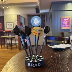 May include: A set of darts in a black and white holder with the name "BRAD" on it. The darts have black and yellow flights, and blue and white shafts. The darts are on a wooden table in a pub setting.