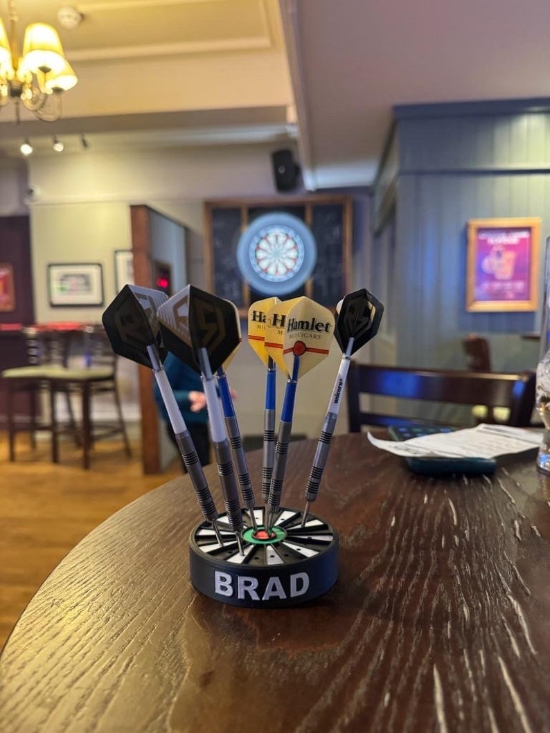 May include: A set of darts in a black and white holder with the name "BRAD" on it. The darts have black and yellow flights, and blue and white shafts. The darts are on a wooden table in a pub setting.