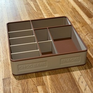 May include: A beige and brown rectangular snack organizer with multiple compartments. The organizer has the words "CHOC BARS" and "SNACKS" embossed on the front. The compartments are designed to hold various types of snacks.