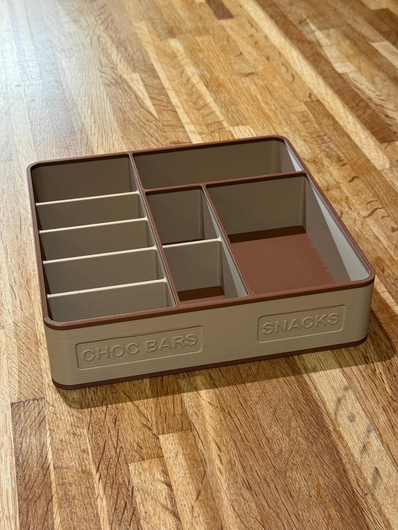 May include: A beige and brown rectangular snack organizer with multiple compartments. The organizer has the words "CHOC BARS" and "SNACKS" embossed on the front. The compartments are designed to hold various types of snacks.
