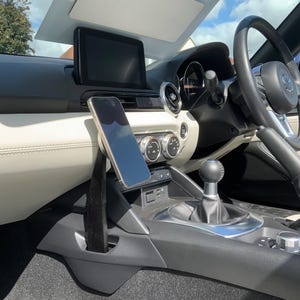 May include: Interior view of a car, showcasing a phone mounted on a black holder. The car's dashboard features a central display, air vents, and a gear shift. The steering wheel and seats are visible, with a bright sky reflected in the windshield.
