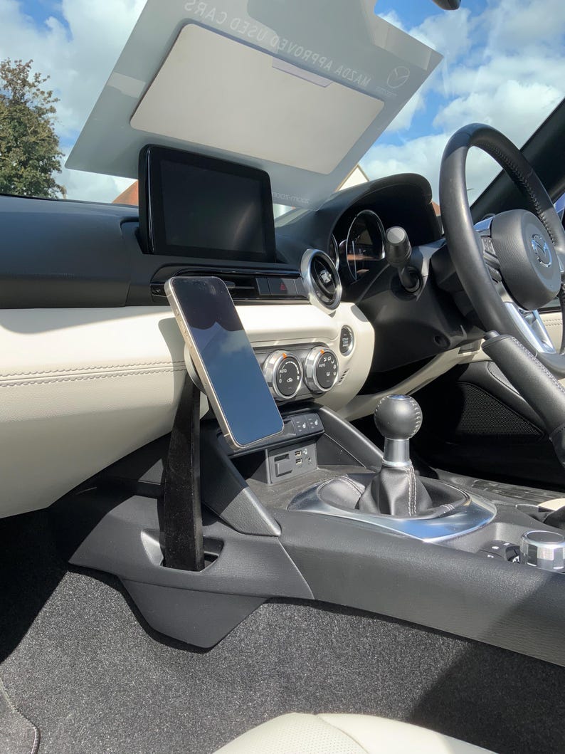May include: Interior view of a car, showcasing a phone mounted on a black holder. The car's dashboard features a central display, air vents, and a gear shift. The steering wheel and seats are visible, with a bright sky reflected in the windshield.
