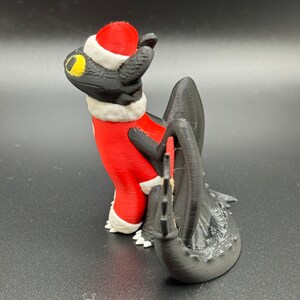 May include: A black and red 3D printed figurine of a dragon wearing a Santa hat and a red and white suit. The dragon is looking up and has a yellow eye.